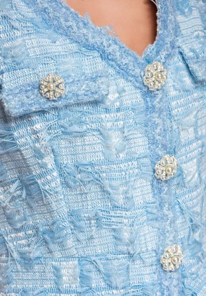 Chaqueta de tela texturizada azul claro con botones en forma de flor de perlas y cristales en el cuello y el frente.