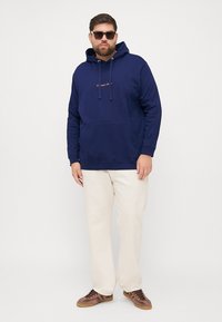 Marineblauwe hoodie met voorzak, capuchon met trekkoord en geborduurd logo. Gecombineerd met crèmekleurige broek en bruine sneakers.