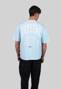 Svetle modré tričko s krátkým rukávem, které má bílý text: "CRYSTAL" a "Nordic Nights Dive into Darkness." Model nosí černé kalhoty.