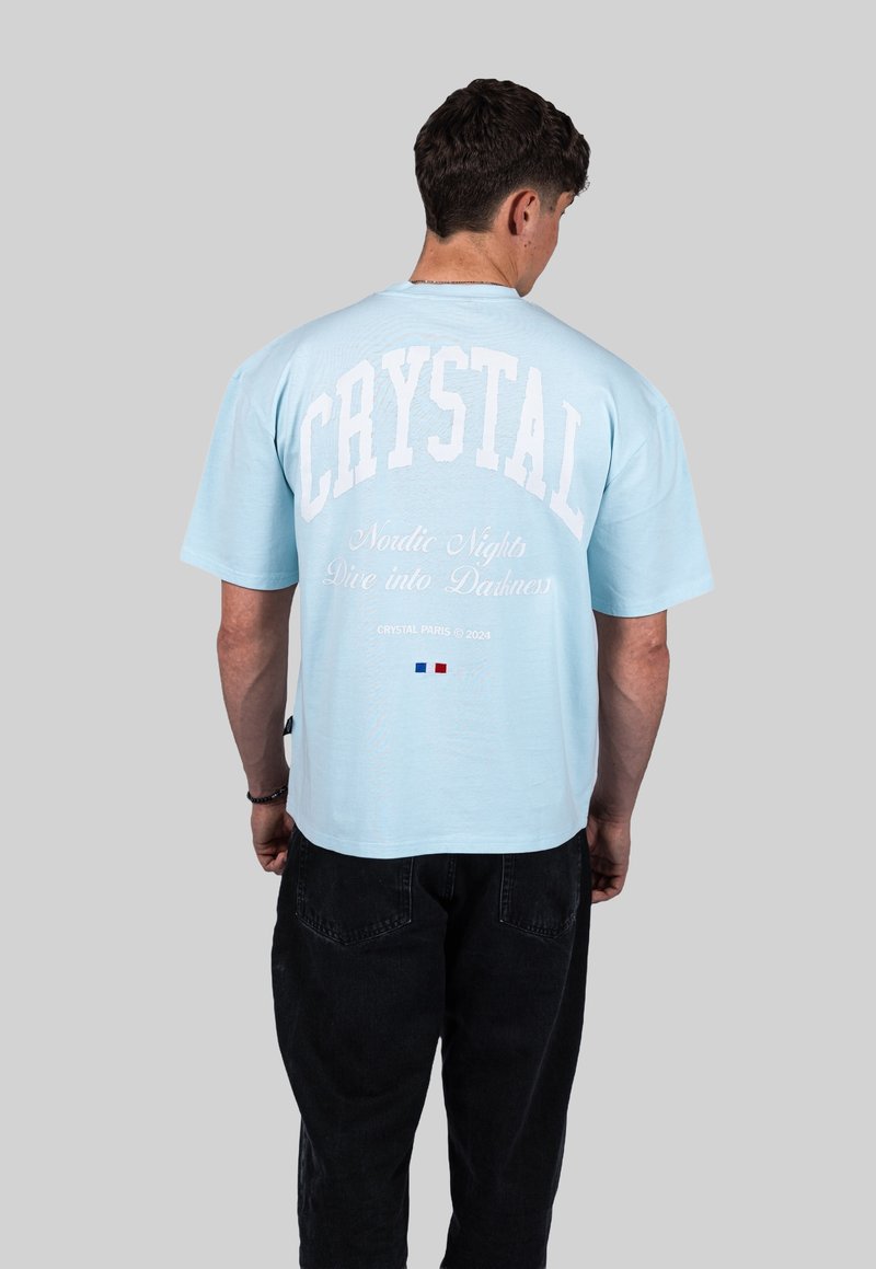 Svetle modré tričko s krátkým rukávem, které má bílý text: "CRYSTAL" a "Nordic Nights Dive into Darkness." Model nosí černé kalhoty.
