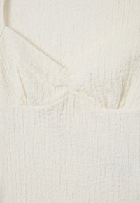 Top de crema claro con una tela texturizada y arrugada. Diseño de escote en V con un detalle anudado gathered en el busto. Textura suave y lisa.