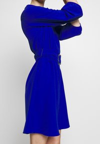 Kungsblå klänning i texturerad tyg, med en figursydd liv, långa ärmar och midja med bälte och metallspänne, utsvängd kjol.