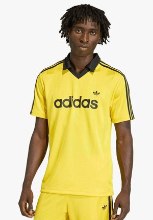 Giovane uomo dalla pelle scura con dreadlocks che indossa una maglia sportiva gialla Adidas e pantaloncini abbinati con logo e strisce nere.