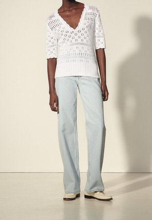 Persona vistiendo un suéter blanco de cuello en V tejido, con mangas hasta el codo, y jeans anchos de tiro alto de color azul claro, combinados con mocasines crema, de pie frente a una pared neutral.