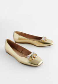 Chaussures plates dorées avec un bout carré, ornées d'un nœud décoratif avec des perles et des accents métalliques. Fini lisse et brillant.