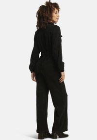 Tuta in velluto a coste nera con maniche lunghe, vita aderente e gambe larghe; presenta un colletto e due tasche posteriori. Abbinata a stivali con tacco.