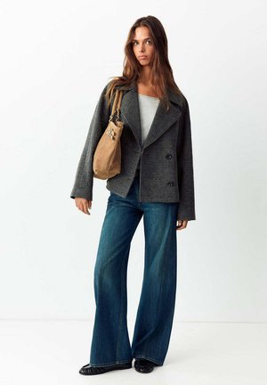 Giovane donna che indossa una giacca grigia a quadri, jeans blu a gamba larga, scarpe nere e una borsa a tracolla color cuoio, in piedi davanti a uno sfondo bianco.