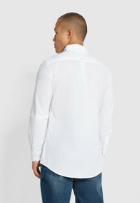 Camicia bianca a maniche lunghe con colletto a punta, orlo curvo e piega sul retro. Tessuto liscio, design aderente e bottoni a contrasto sui polsini.