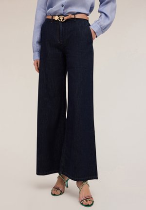 Donkerblauwe wijde jeans met een hoge taille, gecombineerd met een lichtblauw overhemd met knopen en een beige riem met een gouden gesp. Groene en rozenkwarts gekruiste hakken.