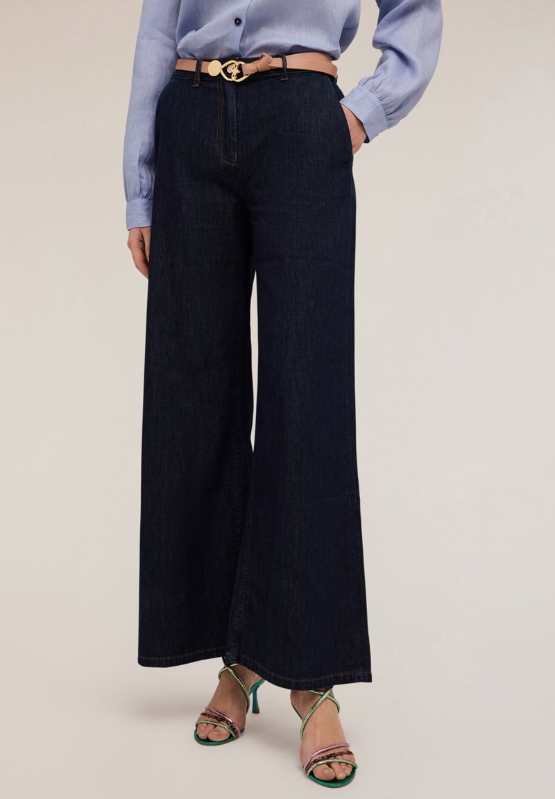 Tummansinisiä leveälahkeisia farkkuja, joissa on korkea vyötärö, yhdessä vaaleansinisen napitettavan paidan ja vaaleanruskean vyön, jossa on kultainen solki, kanssa. Vihreät ja vaaleanpunaiset remmikengät.