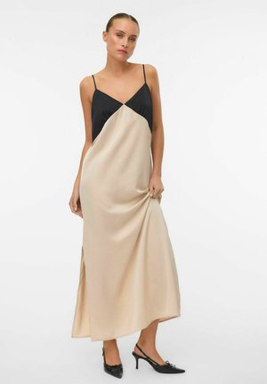 Nő bézs és fekete színű, vékony pántos, ujjatlan maxi ruhában, fekete hegyes orrú slingback magassarkúban, egyszínű fehér háttér előtt áll.