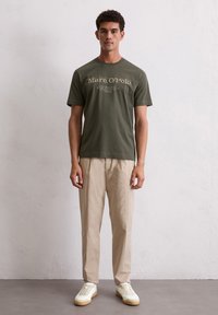 Olijfgroene katoenen t-shirt met korte mouwen, voorzien van een bedrukt logo in lichtbeige. Gepaard met beige broek en witte sneakers.