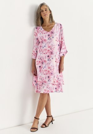 Kvinde med gråt hår iført en lyserød knælang kjole med blomsterprint og sorte sandaler med åben tå, stående op ad en hvid væg.