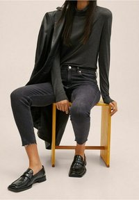 Haut noir à manches longues, jean court en denim foncé avec des ourlets effilochés, et mocassins noirs, assis sur un tabouret en bois devant un fond neutre.