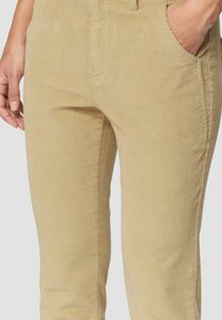 Khakifarbene Cordhosen mit vertikaler Rippstruktur, seitlichen Taschen und tailliertem Schnitt. Sichtbare Nähte betonen die Form und das Design.