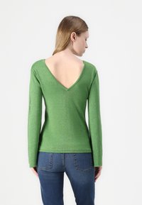Grønn, langermet topp laget av metallisk stoff med v-hals og åpen rygg. Teksturert, tettsittende design, kombinert med mørkeblå jeans.