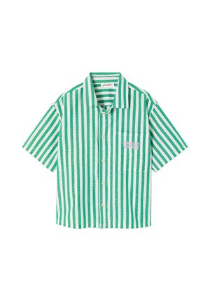 Enfant aux longs cheveux blonds portant une chemise oversize rayée verticale verte et blanche et un short assorti, debout avec des sandales marron.