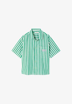 Enfant aux longs cheveux blonds portant une chemise oversize rayée verticale verte et blanche et un short assorti, debout avec des sandales marron.