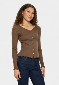 Vrouw met krullend haar, gekleed in een bruine blouse met knopen en donkere spijkerbroek, staand tegen een effen witte achtergrond.