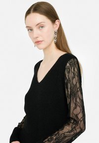 Maglione nero con scollatura a V e corpo in maglia testurizzata, con maniche lunghe in pizzo nero trasparente. Accenti di collegamento notevoli sugli orecchini.