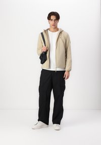 Lettvekts hettejakke i tan over en hvit t-skjorte, kombinert med svarte cargo-bukser og hvite sneakers. Personen holder en liten svart bag.
