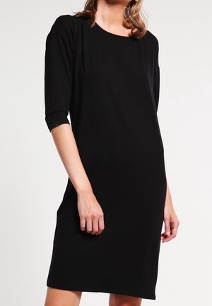 Femme portant une robe noire unie jusqu'aux genoux avec des manches trois-quarts, debout devant un fond clair uni.