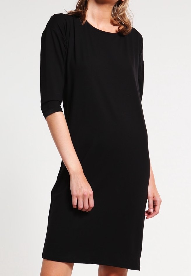 Femme portant une robe noire unie jusqu'aux genoux avec des manches trois-quarts, debout devant un fond clair uni.