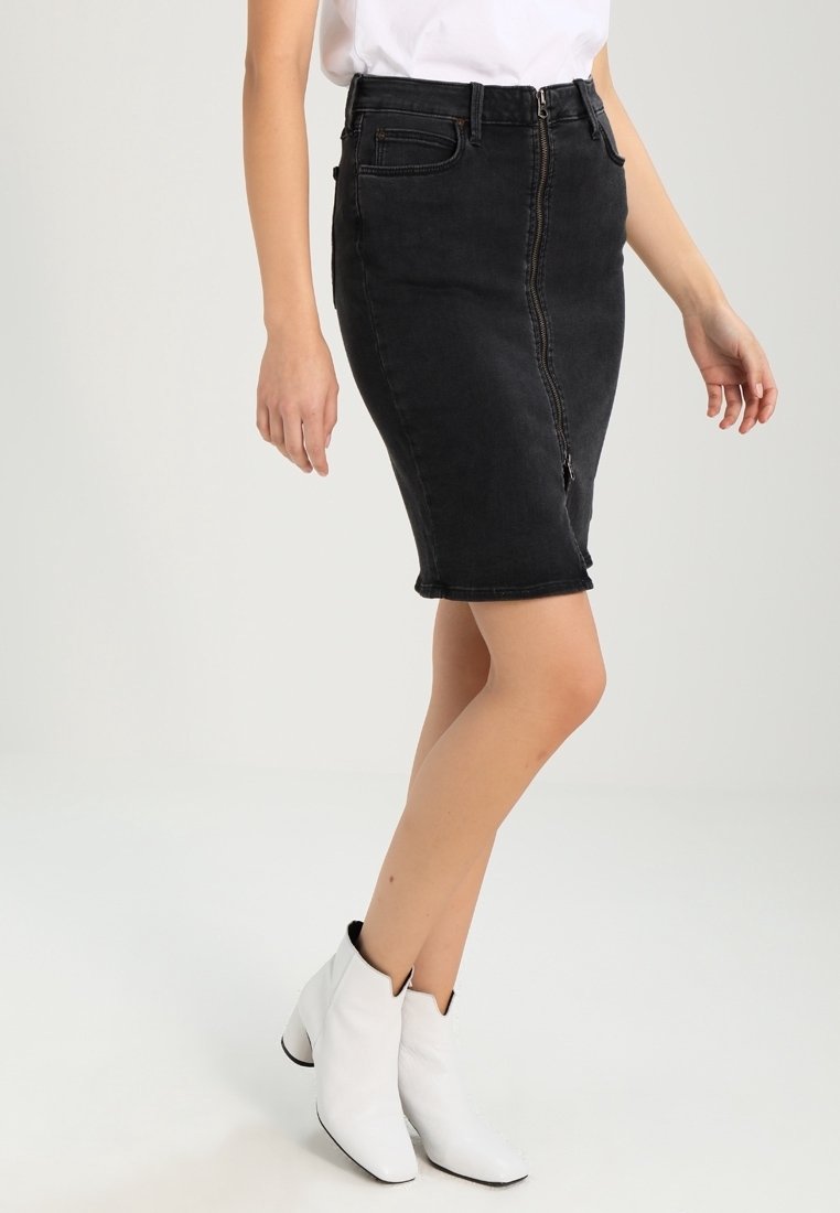 Falda lápiz de mezclilla negra con cremallera delantera y forma ajustada. El modelo lleva botas blancas hasta el tobillo, destacando un look limpio y moderno.