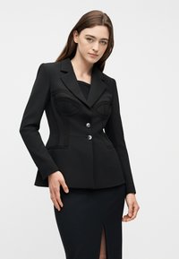Νεαρή γυναίκα που φοράει στενό μαύρο σακάκι με διακριτικές πιέτες στο στήθος και κουμπιά, συνδυασμένο με μαύρη φούστα που έχει σκίσιμο μπροστά.