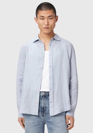 Mann trägt ein hellblaues Hemd über einem weißen Tanktop und ausgewaschenen blauen Jeans, steht vor einem einfarbigen Hintergrund.