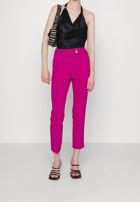 Vrouw in een zwarte haltertop, felle roze slim-fit broek met knopen, zwarte sleehakken en een schoudertas met zebra print tegen een witte achtergrond.