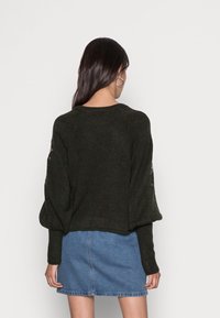Pull en maille vert foncé avec des manches oversized, orné d'accents brodés sur les épaules. Accordé avec une jupe en denim.
