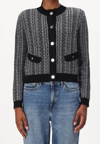 Svart och vit mönstrad kofta med rund halsringning, knäppning med knappar, två framfickor och ribbad ärm. Matchad med ljusblå jeans.