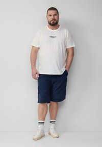 Biała bawełniana koszulka z logo, granatowe szorty, białe trampki i pasiaste skarpetki. Mężczyzna stoi, ręce w kieszeniach, neutralne tło.