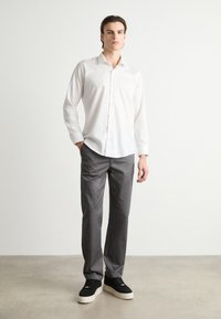 Vit långärmad skjorta med knappnerkoll, i kombination med gråbyxor. Modellen bär svarta sneakers med vita sulor. Enkelt, rent design.