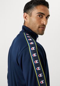 Veste de survêtement zippée marine avec accents de logo en rouge et blanc le long des manches, bordée d'une bande jaune, texture de tissu lisse.