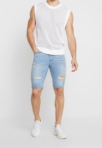 Top de malla sin mangas blanco con dobladillo acanalado; shorts de mezclilla azul claro con detalles desgastados; usado con zapatillas blancas.