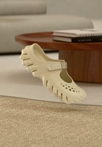 Chaussure Crocs beige avec dessus perforé et semelle épaisse, positionnée en suspension près d'une table basse en bois avec des livres empilés.