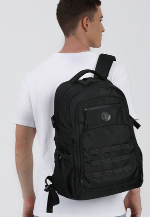 Uomo con maglietta bianca che indossa uno zaino nero grande con molte tasche con cerniera e tasche laterali, visto di spalle.