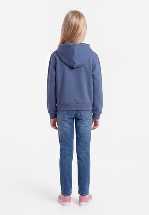 Enfant aux longs cheveux blonds portant un sweat à capuche bleu, un jean bleu et des baskets roses, debout de dos sur un fond blanc.