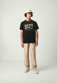 Nuori mies, jolla on päällään musta "Jack & Jones" -t-paita, vaaleat housut, valkoiset lenkkarit ja vaaleanbeige baseball-lippalakki, seisoo yksinkertaista taustaa vasten.