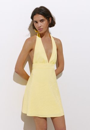 Femme aux cheveux courts et bruns portant une robe jaune sans manches à col halter, debout contre un mur blanc.