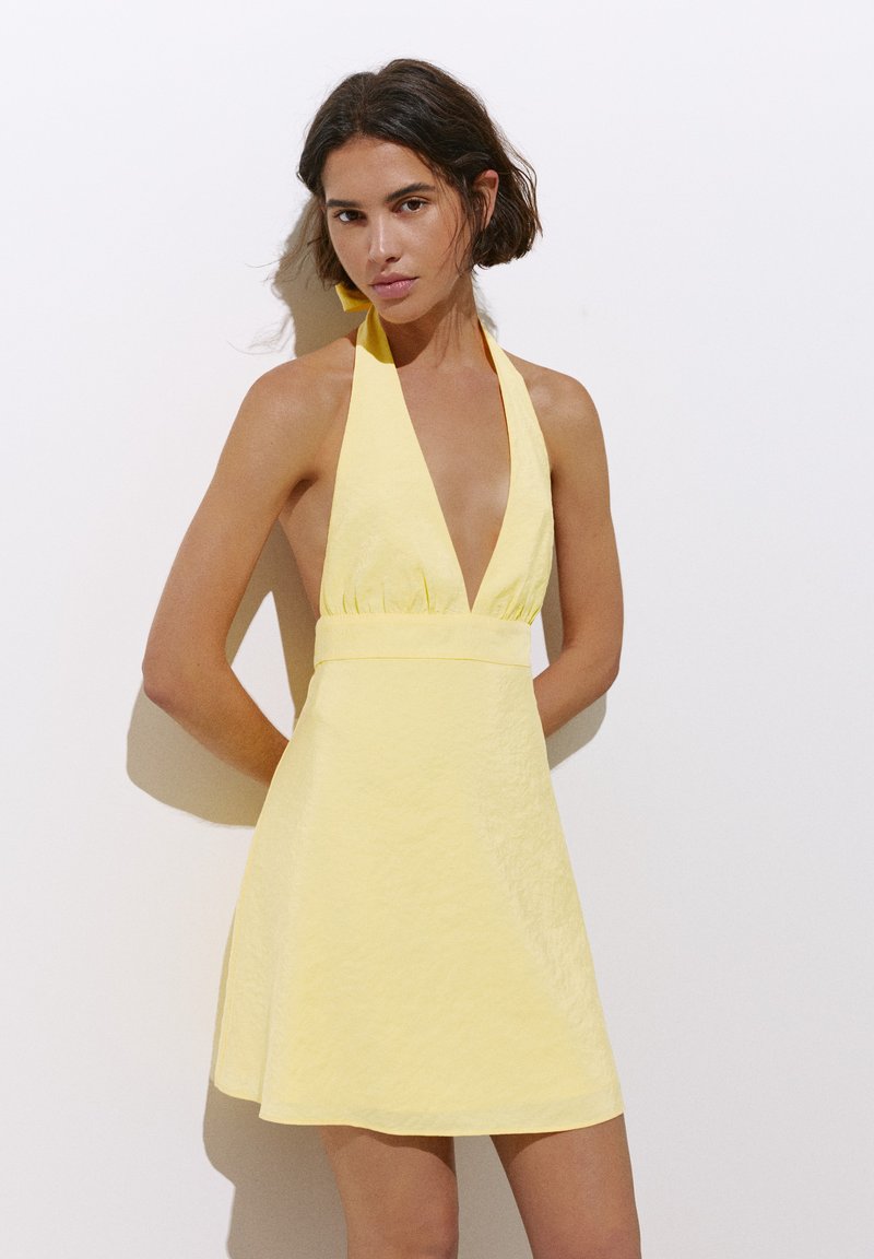 Femme aux cheveux courts et bruns portant une robe jaune sans manches à col halter, debout contre un mur blanc.