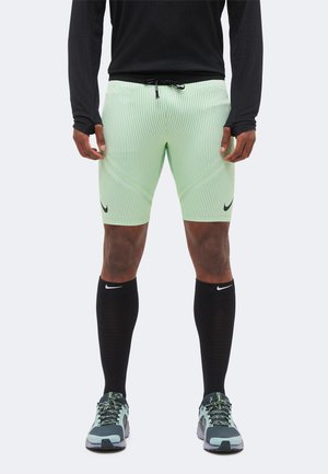 Homme portant un short de sport rayé vert clair, un haut noir à manches longues, des chaussettes noires montantes et des chaussures de course vertes et noires.