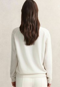 Pull crème avec un tissu doux et texturé. Caractérisé par une coupe décontractée, des manches longues et des poignets côtelés, porté sur un pantalon clair.