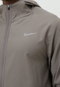 Gros plan d'une personne portant une veste Nike taupe à fermeture éclair avec capuche et logo Nike Swoosh réfléchissant sur la poitrine.