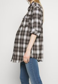 Camisa de cuadros en negro, blanco y marrón, con dos bolsillos en el pecho, mangas enrolladas y un corte holgado. Dobladillo trasero más largo con un corte recto.