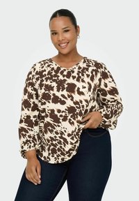 Blandena bluzica v brown in krem vzorcu z V-izrezom in dolgimi rokavi, ki ima nabran rob in ohlapen kroj. V kombinaciji s temno modrimi džinsami.