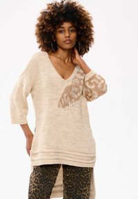 Gehaakte beige trui met een diepe V-hals en driekwart mouwen, voorzien van een bruine dierenprintaccent aan de linkerkant. Getextureerde stof.