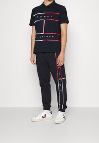 Laivastonsininen polo-paita, jossa on valkoisia ja punaisia graafisia viivoja ja logo; yhdistettynä vastaaviin verkkareihin, joissa on sivuviiva ja logo. Valkoiset tennarit.